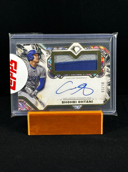 A Shohei Ohtani baseball trading card in a clear protective case, displayed on a small wooden stand against a black background. The card features a blue fabric swatch, a blue autograph, and a red circular topps sticker on the left.