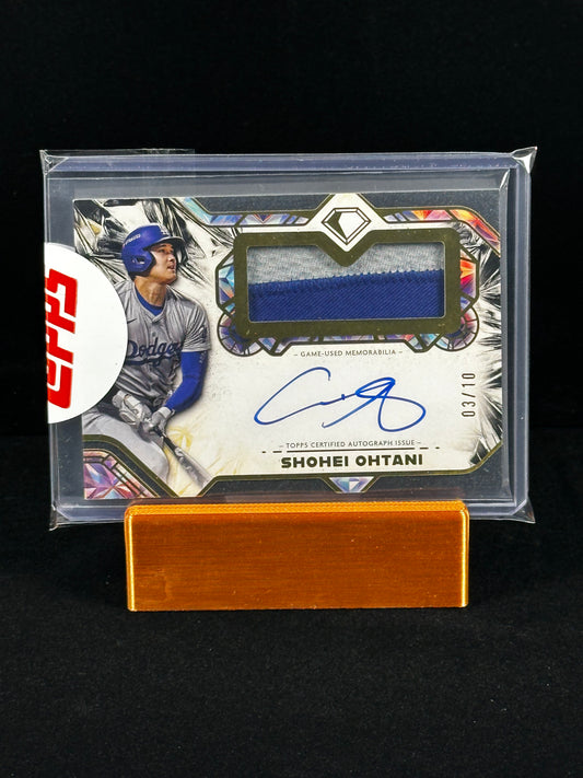 A Shohei Ohtani baseball trading card in a clear protective case, displayed on a small wooden stand against a black background. The card features a blue fabric swatch, a blue autograph, and a red circular topps sticker on the left.