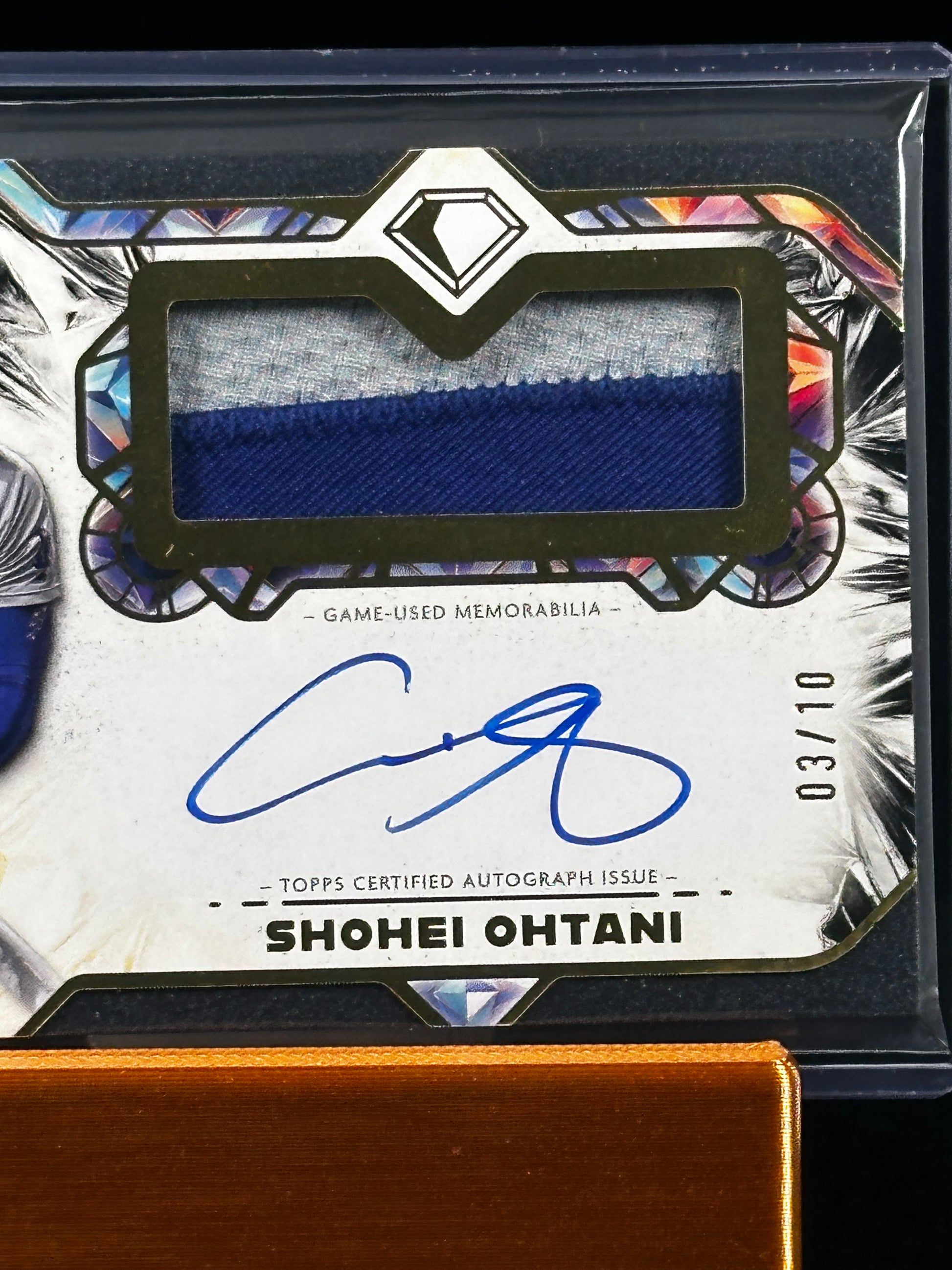 A close-up of a collectible sports memorabilia card featuring a blue fabric swatch, an autographed signature, and the name Shohei Ohtani, displayed on a reflective surface.