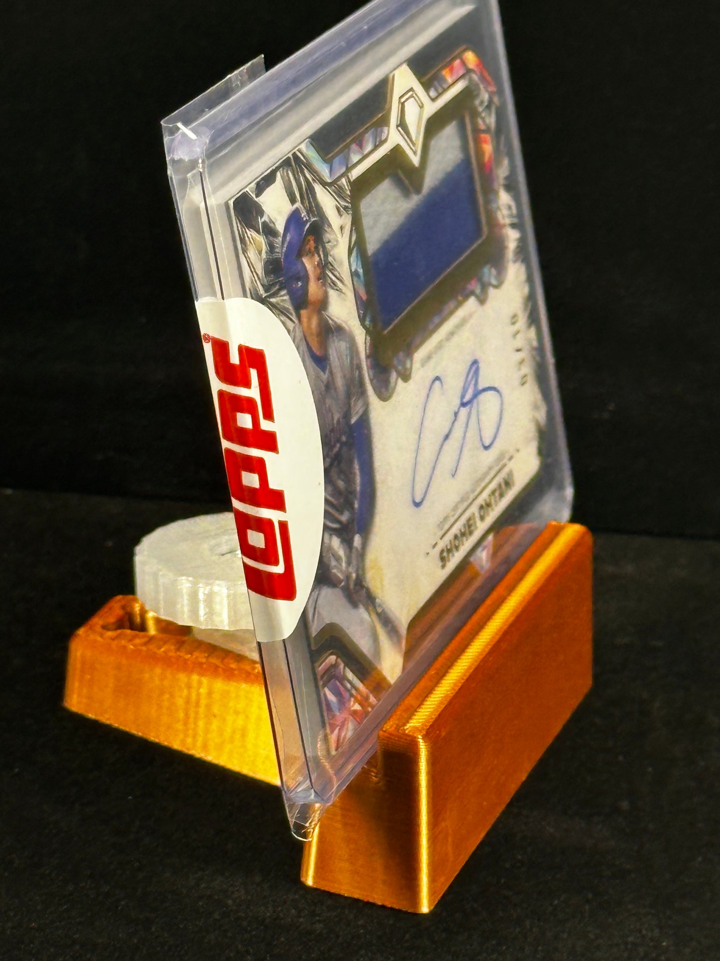 A signed baseball card in a clear protective case, displayed on a gold stand against a black background. The card features a blue fabric swatch and a blue autograph, with a white circular topps sticker seal