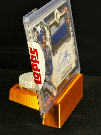 A signed baseball card in a clear protective case, displayed on a gold stand against a black background. The card features a blue fabric swatch and a blue autograph, with a white circular topps sticker seal