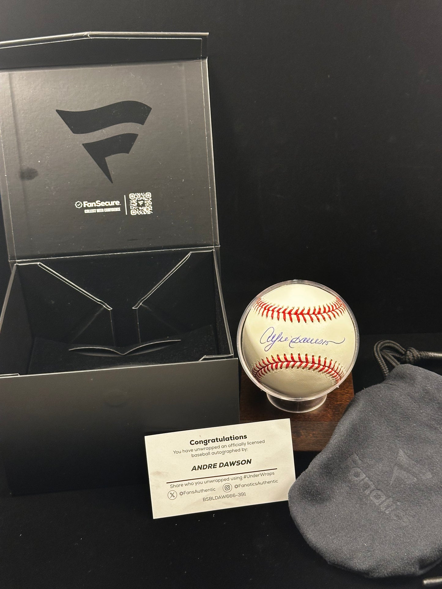 A signed Andre Dawson baseball displayed in a clear stand, next to its black FanSecure presentation box, a white congratulations card, and a gray drawstring pouch on a black background.