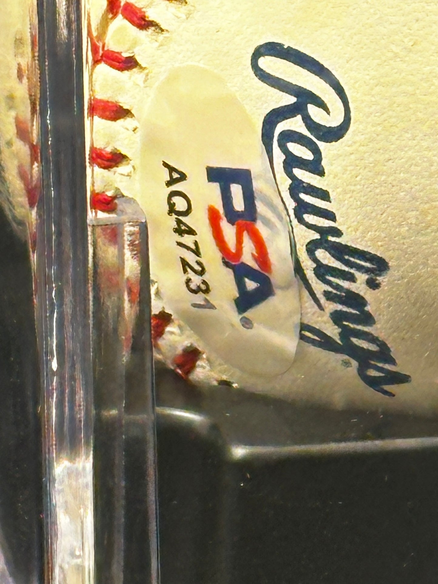 A close-up of a white baseball with red stitching, featuring a PSA authentication sticker and a blue Rawlings logo, partially visible behind a clear display case.