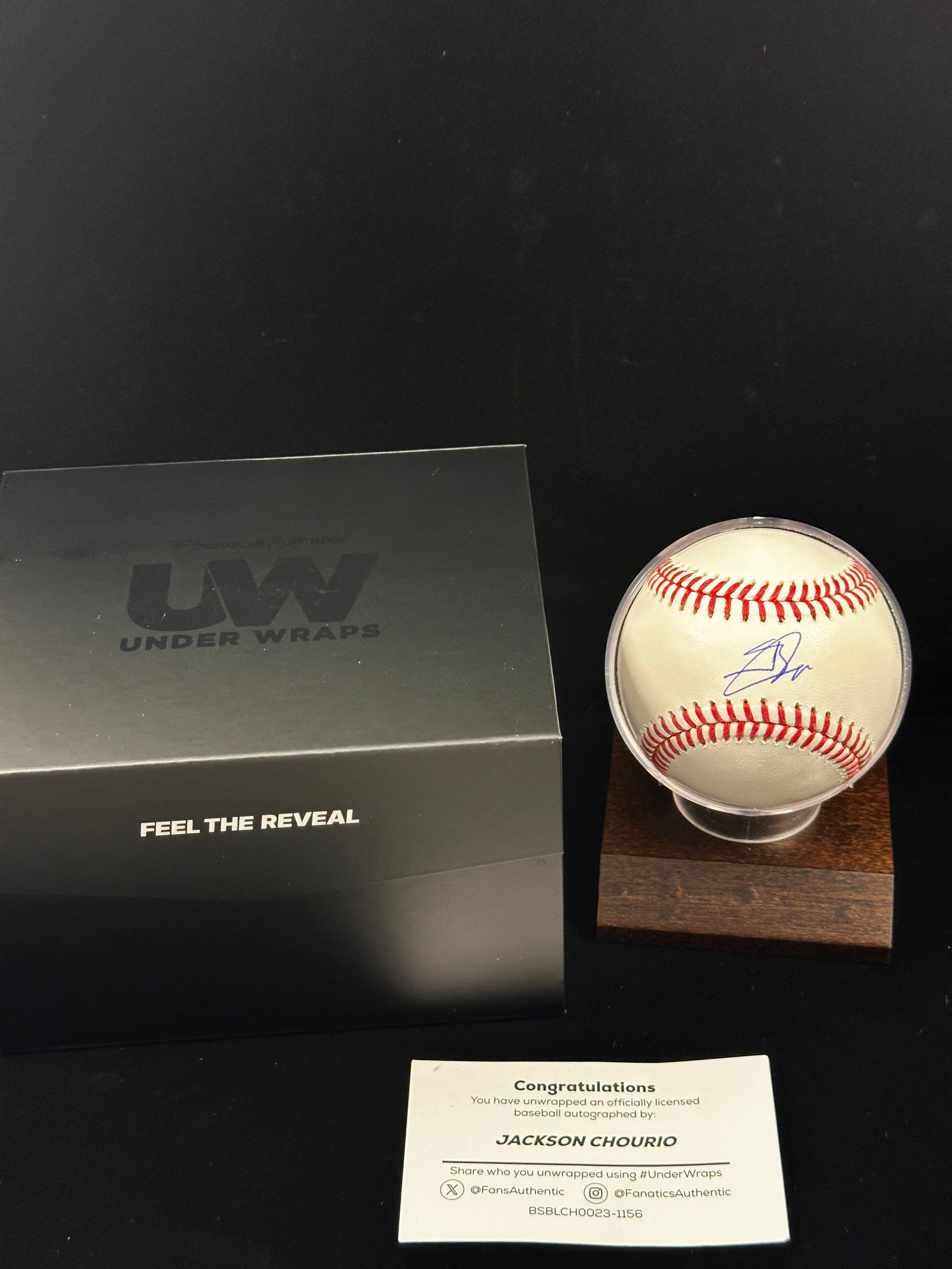 A Jackson Chourio autographed baseball displayed in a clear sphere on a wooden stand, alongside a black Fanatics Under Wraps presentation box and a small white congratulations card on a black background.