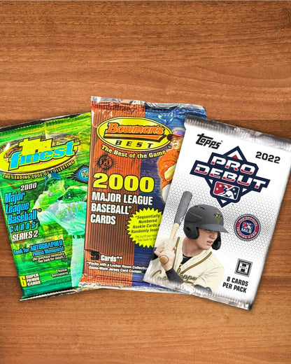 Three baseball trading card booster packs arranged on a wooden surface: a green 2000 Majestic pack, a blue and orange 2000 Bowman Series 2 pack, and a white 2022 Topps Pro Debut pack.