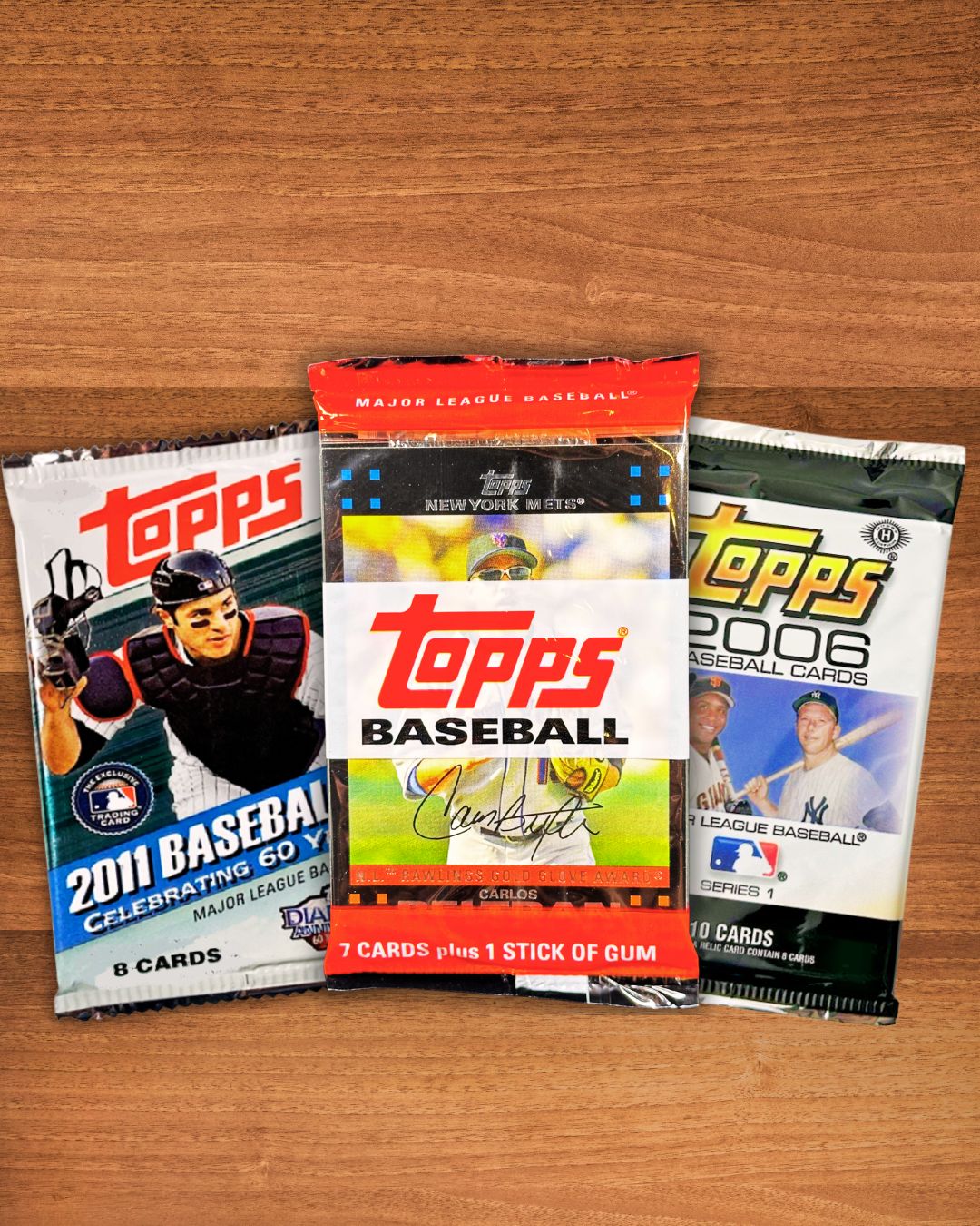 Three Topps Baseball trading card booster packs arranged on a wooden surface: a 2011 baseball pack on the left, a 2006 Topps pack in the center, and a 2006 MLB Baseball pack on the right.