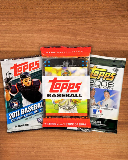 Three Topps Baseball trading card booster packs arranged on a wooden surface: a 2011 baseball pack on the left, a 2006 Topps pack in the center, and a 2006 MLB Baseball pack on the right.