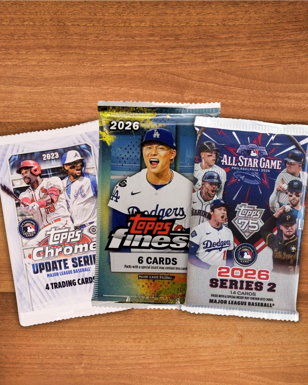 Three Topps baseball trading card packs arranged on a wooden surface: a 2023 Chrome Update Series pack, a 2026 Topps Fines 6-card foil pack, and a 2026 Series 2 All Star Game pack.
