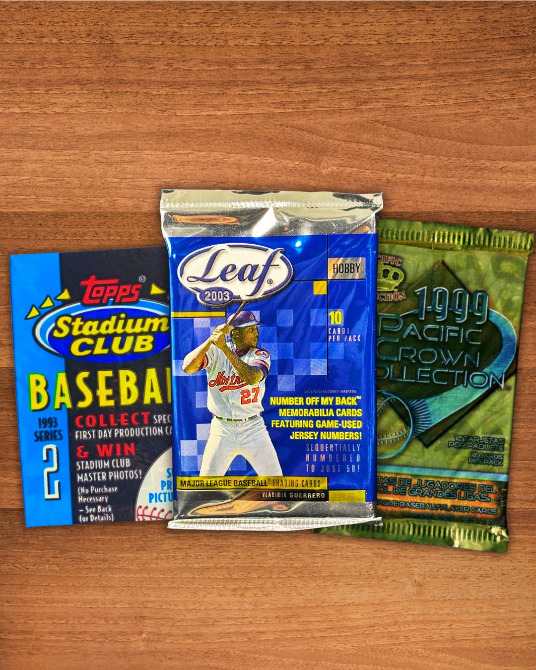Three collectible trading card packs arranged on a wooden tabletop: a blue 1993 Topps Stadium Club baseball pack on the left, a silver and blue Leaf 2003 baseball pack in the center, and a green 1999 Pacific Coast Collection baseball pack on the right.