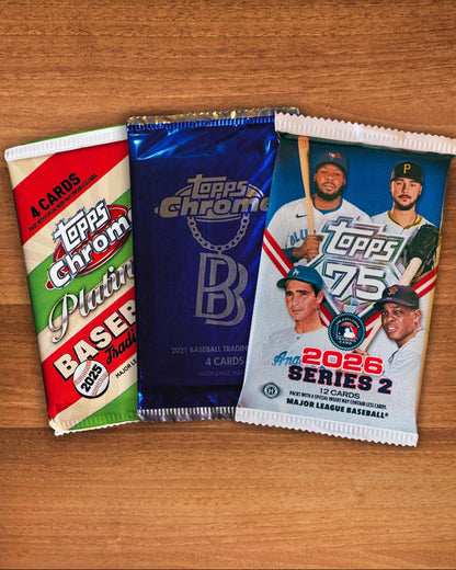 Three Topps-branded baseball trading card booster packs arranged side by side on a wooden surface. The packs include a red, green, and cream 2025 Platinum Baser pack, a metallic blue 2026 Topps Chrome pack, and a white 2026 Series 2 Major League Baseball pack with player photos.
