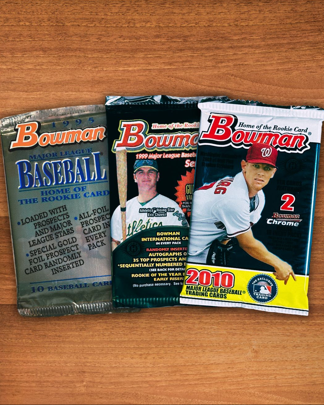Three Bowman baseball trading card booster packs arranged side by side on a wooden tabletop. The packs feature different color schemes, including blue, black, and a white/yellow/red 2010 edition, with player photos and Bowman branding visible.