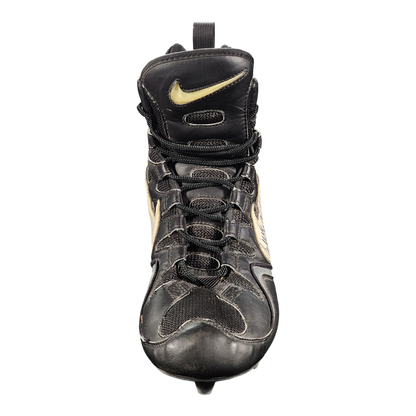 A black Nike high-top baseball cleat with gold accents, featuring a leather and mesh upper, lace-up front, and a spike on the sole, shown against a plain white background.